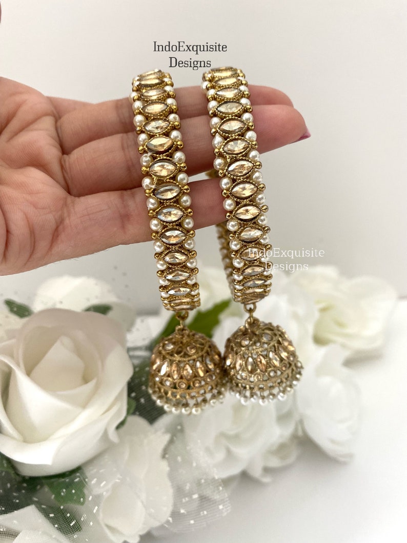 May include: A pair of gold bangles with white pearl accents and dangling gold earrings with white pearl accents. The bangles and earrings are decorated with clear stones.