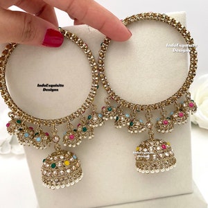 May include: A pair of gold bangles with dangling charms. The charms are decorated with colourful stones and pearls. The bangles are adorned with the text "IndoExquisite Designs".
