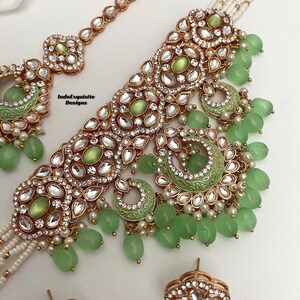 May include: A gold and green Indian bridal necklace set with a choker, a maang tikka, and earrings. The necklace features a multi-layered design with green stones and pearls. The maang tikka is a delicate chain with a pendant. The earrings are round with green stones and pearls.
