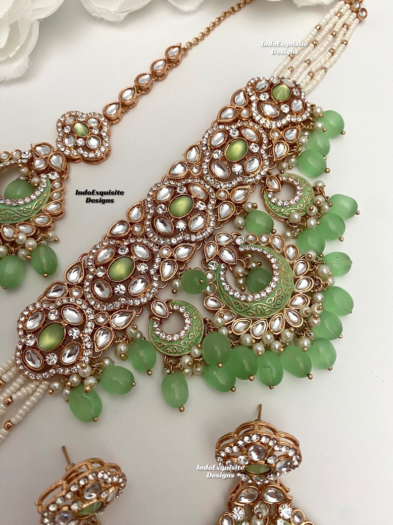 May include: A gold and green Indian bridal necklace set with a choker, a maang tikka, and earrings. The necklace features a multi-layered design with green stones and pearls. The maang tikka is a delicate chain with a pendant. The earrings are round with green stones and pearls.