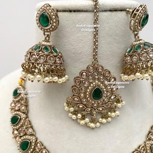 May include: An antique gold-toned jewelry set featuring a necklace, earrings, and a headpiece. The set is adorned with green gemstones, small pearls, and clear crystals. The earrings are a jhumka style. The text "IndoExquisite Designs" is visible.