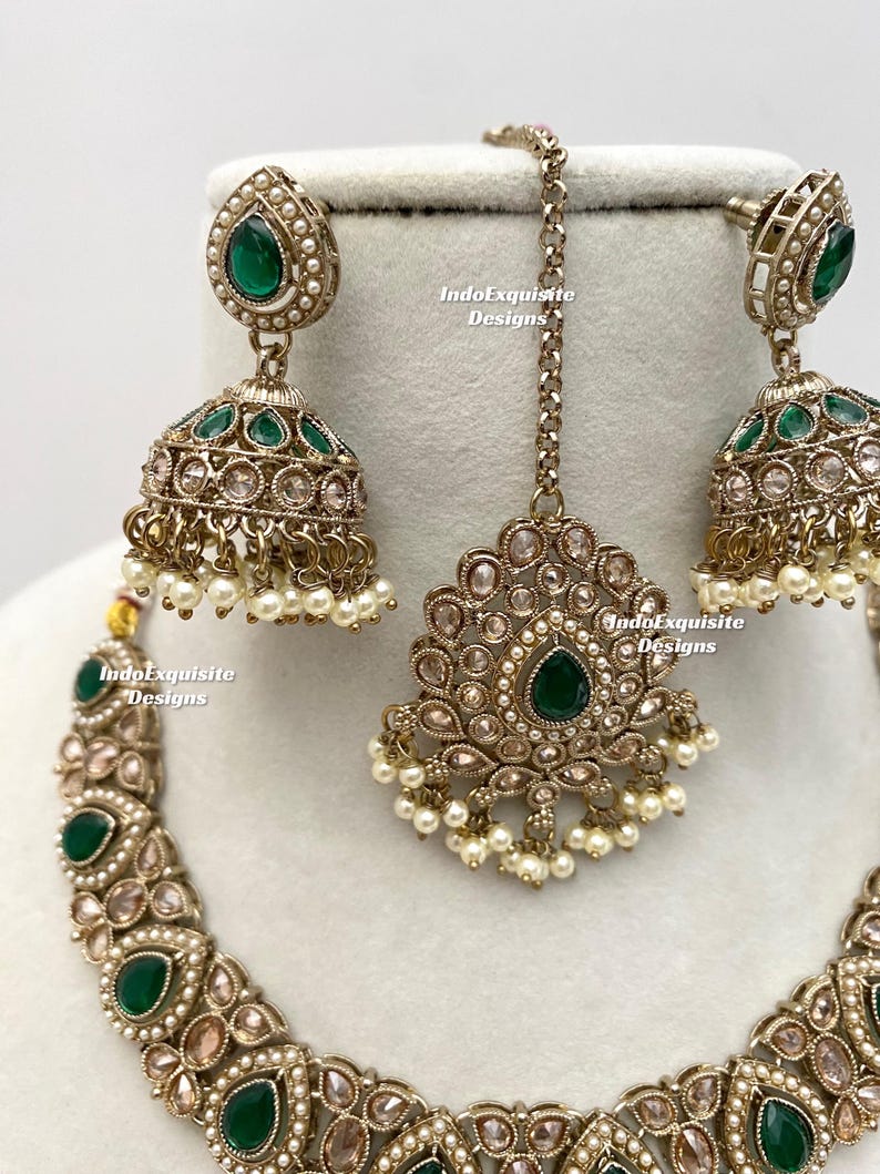 May include: An antique gold-toned jewelry set featuring a necklace, earrings, and a headpiece. The set is adorned with green gemstones, small pearls, and clear crystals. The earrings are a jhumka style. The text "IndoExquisite Designs" is visible.
