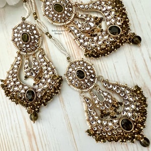 May include: A pair of gold-tone earrings with green and white stones. The earrings are made of metal and have a traditional Indian design. The earrings are hanging from a chain with white beads.