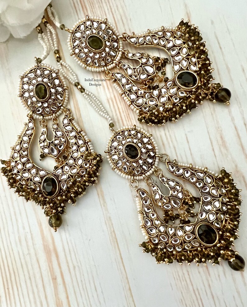 May include: A pair of gold-tone earrings with green and white stones. The earrings are made of metal and have a traditional Indian design. The earrings are hanging from a chain with white beads.
