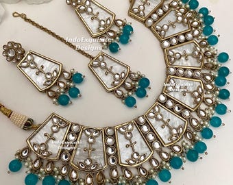 Puede incluir: Un conjunto de joyería que incluye un collar, dos pendientes y un maang tikka. Cada pieza tiene marcos de metal dorado, incrustaciones de nácar blanco con motivos florales y cuentas turquesas colgantes. El texto IndoExquisite Designs es visible.