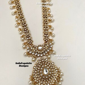 May include: A long gold necklace with white pearls and a large, ornate pendant. The pendant is made of gold and has a teardrop-shaped stone in the center. The necklace is decorated with small, white stones.