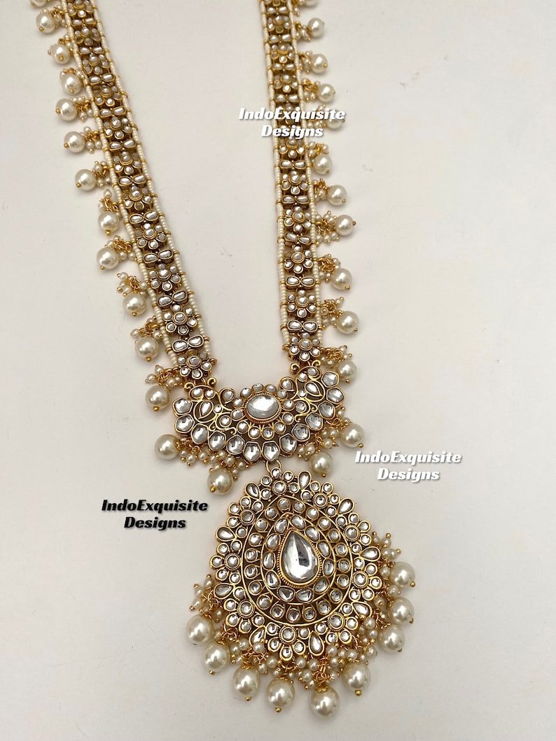 May include: A long gold necklace with white pearls and a large, ornate pendant. The pendant is made of gold and has a teardrop-shaped stone in the center. The necklace is decorated with small, white stones.
