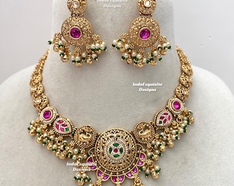 Puede incluir: Un conjunto de collar y pendientes en tono dorado con intrincados trabajos en metal, motivos circulares y piedras incrustadas de color rosa y verde. La joyería está adornada con pequeñas cuentas blancas tipo perla y acentos verdes, con el texto IndoExquisite Designs.