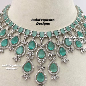 May include: A silver necklace with a large, teardrop-shaped, light green stone pendant surrounded by smaller clear stones. The necklace has a row of rectangular, light green stones at the top and a delicate, floral design.