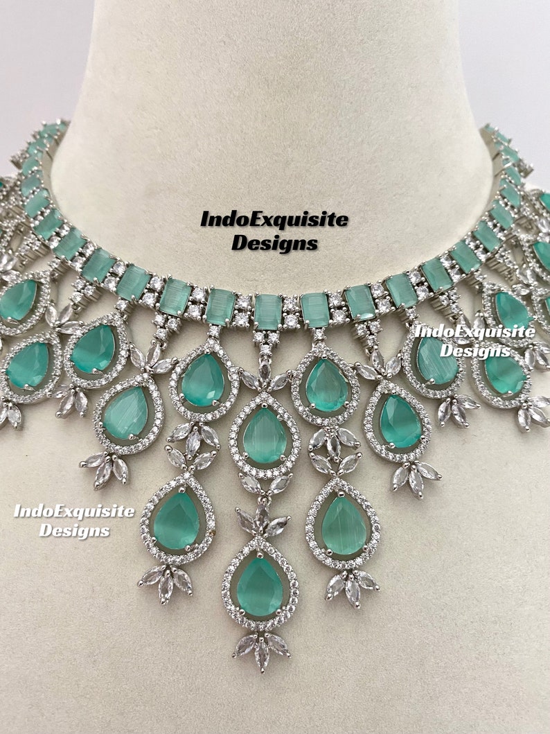 May include: A silver necklace with a large, teardrop-shaped, light green stone pendant surrounded by smaller clear stones. The necklace has a row of rectangular, light green stones at the top and a delicate, floral design.