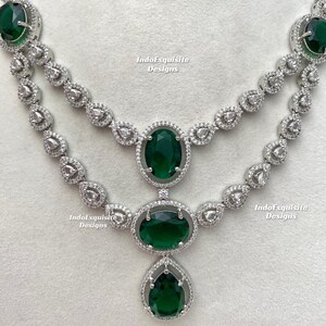 May include: A two-strand necklace and matching earrings set. The jewelry features large, oval, emerald-green gemstones surrounded by a halo of smaller, clear stones. The necklace has a silver-colored metal setting and a teardrop-shaped pendant.