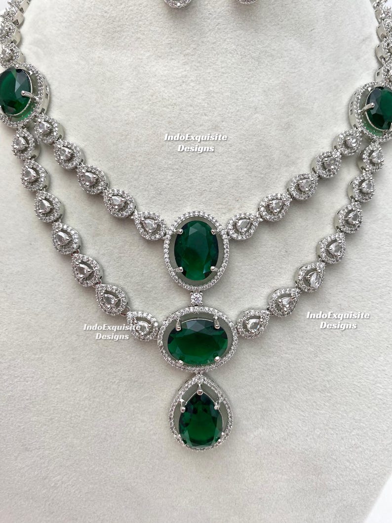 May include: A two-strand necklace and matching earrings set. The jewelry features large, oval, emerald-green gemstones surrounded by a halo of smaller, clear stones. The necklace has a silver-colored metal setting and a teardrop-shaped pendant.