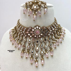 May include: A gold and silver choker necklace with pink and white beads. The necklace has a large, ornate centerpiece with a pink mirror. The necklace is on a white background. IndoExquisite Designs is written on the background.