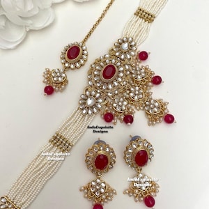 May include: A gold and red Indian bridal jewellery set featuring a choker necklace, earrings, and a maang tikka. The necklace is made of pearls and has a large, ornate pendant with red stones. The earrings are also ornate and feature red stones. The maang tikka is a small, delicate piece with a red stone.