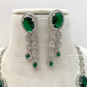 May include: A silver-toned jewelry set featuring oval-cut emerald green gemstones and clear, sparkling accents. The earrings have a cascading design with teardrop-shaped green gems. The necklace is partially visible, with a matching oval green stone.