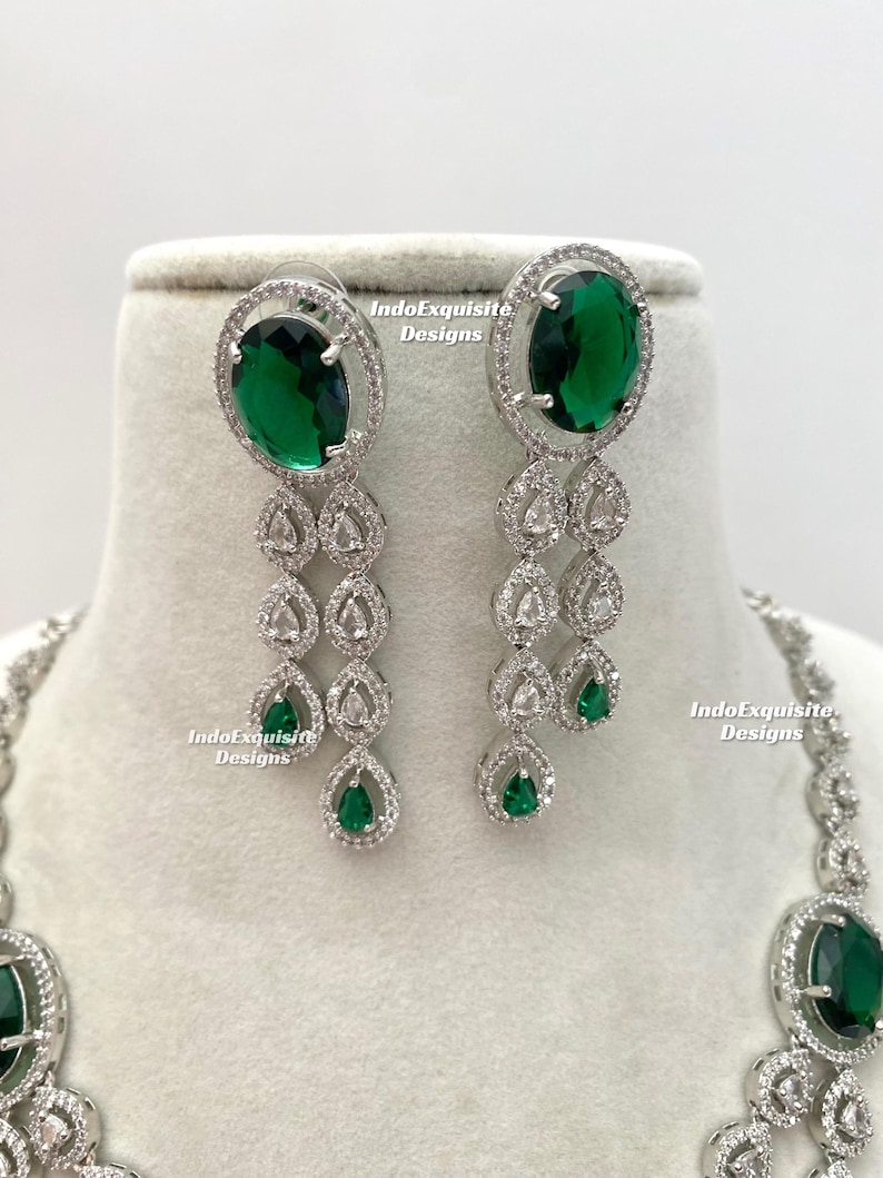 May include: A silver-toned jewelry set featuring oval-cut emerald green gemstones and clear, sparkling accents. The earrings have a cascading design with teardrop-shaped green gems. The necklace is partially visible, with a matching oval green stone.