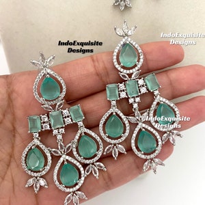 May include: A pair of silver dangle earrings with green teardrop-shaped stones and clear cubic zirconia accents. The earrings feature a three-tiered design with a square-shaped stone on the top tier.