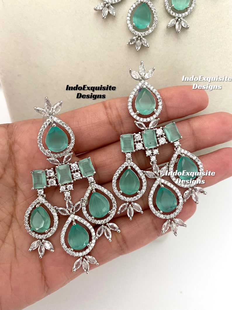 May include: A pair of silver dangle earrings with green teardrop-shaped stones and clear cubic zirconia accents. The earrings feature a three-tiered design with a square-shaped stone on the top tier.