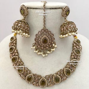 May include: An antique gold-toned jewelry set featuring a necklace, earrings, and a headpiece. The necklace and earrings have olive-green and clear teardrop-shaped stones, with small white pearls. The headpiece is a matching design. The text "IndoExquisite Designs" is visible.