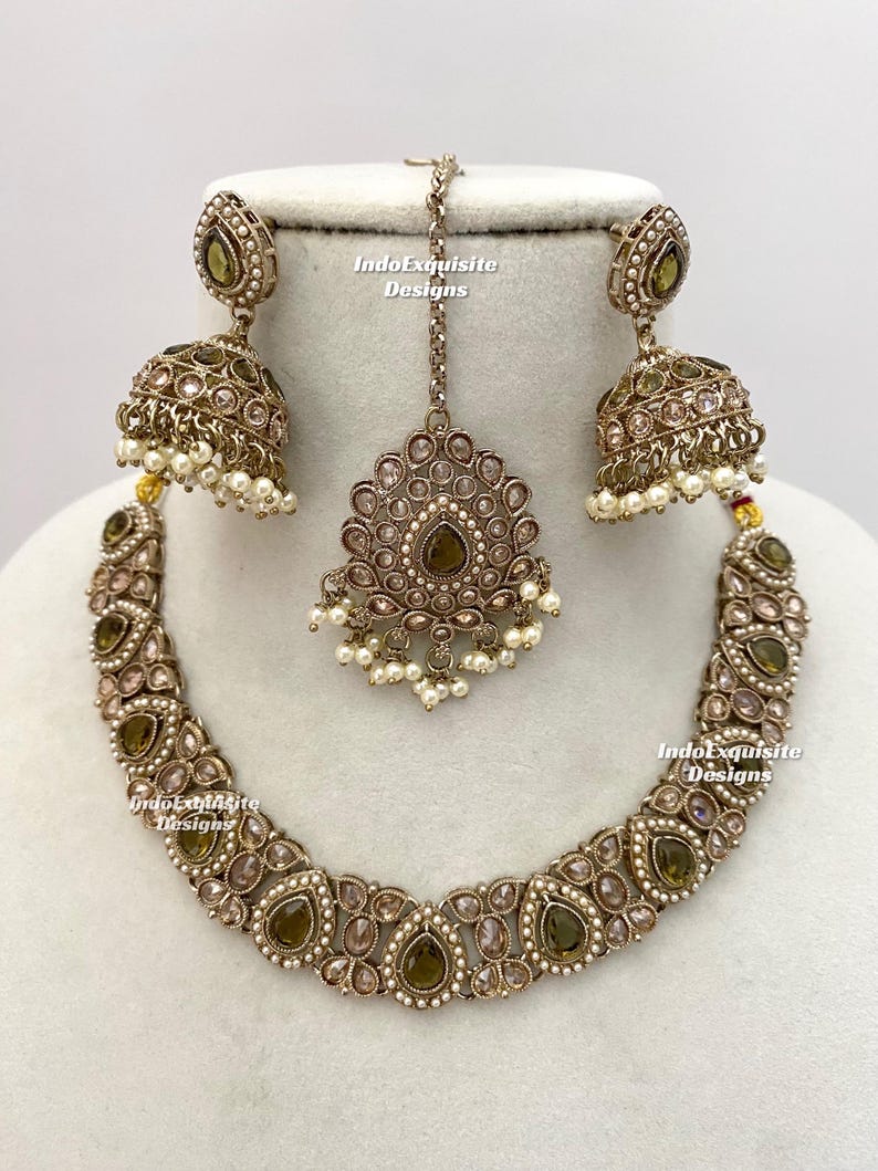 May include: An antique gold-toned jewelry set featuring a necklace, earrings, and a headpiece. The necklace and earrings have olive-green and clear teardrop-shaped stones, with small white pearls. The headpiece is a matching design. The text "IndoExquisite Designs" is visible.