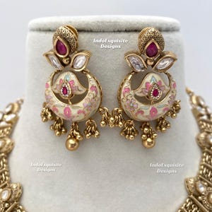 Pu&ograve; includere: Orecchini color oro con un design floreale. Gli orecchini presentano una base color crema con accenti floreali rosa e blu. Ogni orecchino ha una gemma rossa e piccole perline dorate che pendono dal basso.