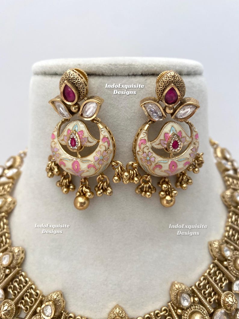 Pu&ograve; includere: Orecchini color oro con un design floreale. Gli orecchini presentano una base color crema con accenti floreali rosa e blu. Ogni orecchino ha una gemma rossa e piccole perline dorate che pendono dal basso.