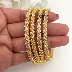 May include: Set of four gold bangles with a row of clear cubic zirconia stones around each bangle. The bangles are stacked on top of each other. The text "IndoExquisite Designs" is visible on the image.