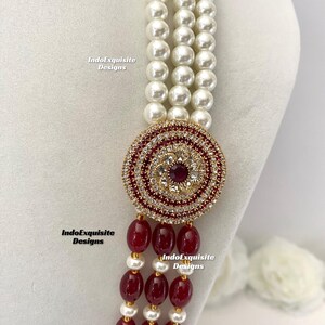May include: A multi-strand necklace featuring white faux pearls and a circular pendant. The pendant is adorned with red and clear crystals, and a central red gemstone. The necklace also includes strands of red beads and gold accents. The text "IndoExquisite Designs" is visible.