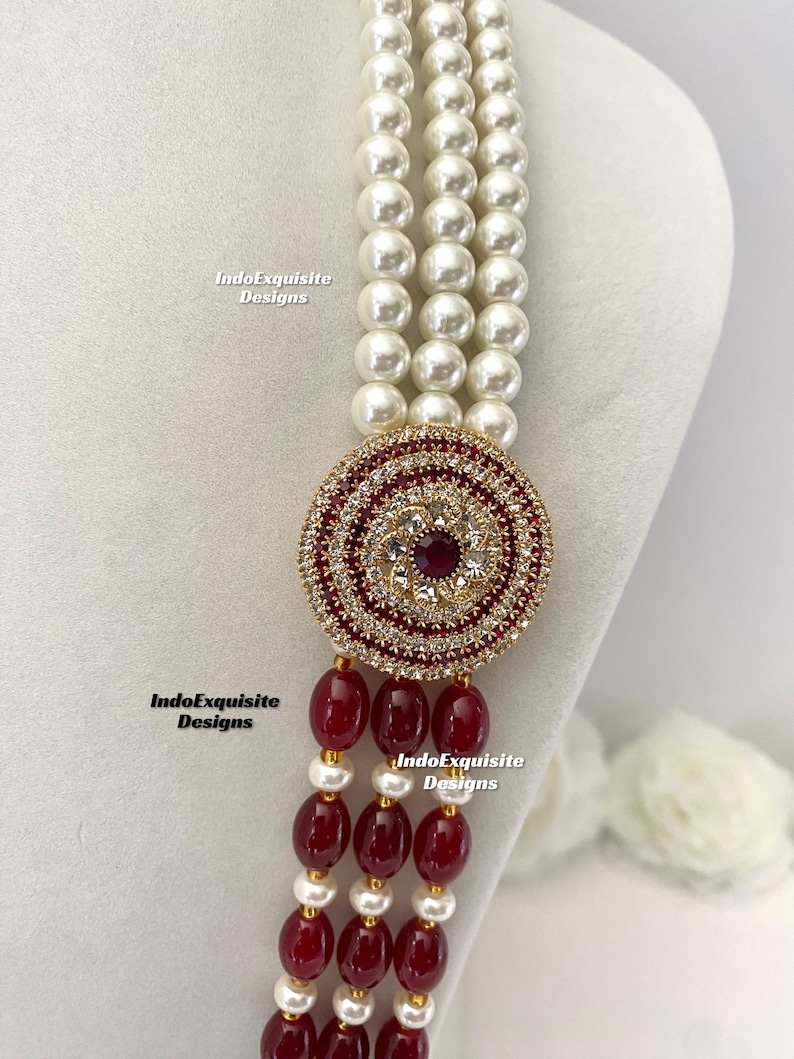 May include: A multi-strand necklace featuring white faux pearls and a circular pendant. The pendant is adorned with red and clear crystals, and a central red gemstone. The necklace also includes strands of red beads and gold accents. The text "IndoExquisite Designs" is visible.