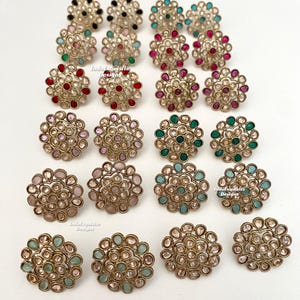 May include: A set of 18 gold-tone stud earrings featuring various coloured stones. The earrings have a circular design with a central stone surrounded by smaller stones. The stones are in shades of pink, red, green, blue, and black.