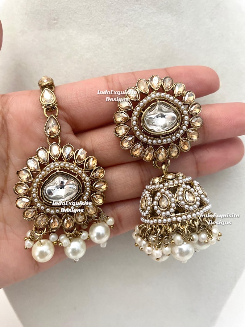 May include: Gold-toned earrings with a floral design, featuring clear and champagne-colored stones, and white pearl accents. The earrings have a dangling bell shape with multiple pearl drops.