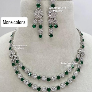 Puede incluir: Un conjunto de collar de dos hilos y pendientes a juego. La joyería presenta un metal de color plateado con piedras transparentes y verde esmeralda. Los pendientes son colgantes y el collar se exhibe en un maniquí. El texto "More colors" es visible.