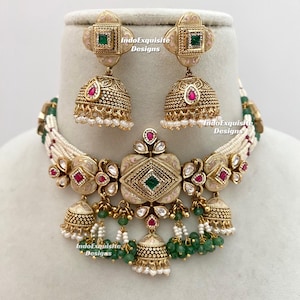 Op de afbeelding: Een sierlijke, goudkleurige ketting en oorbellenset. De ketting heeft een centrale hanger met een groene edelsteen, omringd door rode edelstenen en hangende groene kralen. De tekst "IndoExquisite Designs" is zichtbaar.
