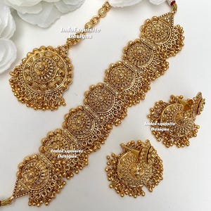 Peut inclure: Un ensemble de bijoux de mariée indien en or, comprenant un collier, des boucles d'oreilles et un maang tikka assortis. Le collier est composé d'une chaîne délicate avec un grand pendentif orné et plusieurs petits pendentifs. Les boucles d'oreilles sont également ornées et présentent un grand pendentif rond. Le maang tikka est une petite pièce ornée qui se porte dans les cheveux.