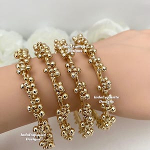 Könnte beinhalten: Vier goldfarbene Armreifen mit floralem Design werden an einem Handgelenk gezeigt. Jeder Armreif ist mit kleinen, runden Perlen verziert, die zu einem blütenartigen Muster angeordnet sind. Der Text "IndoExquisite Designs" ist auf jedem Armreif sichtbar.