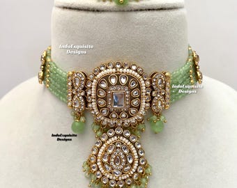 Op de afbeelding: Een groene en gouden choker ketting met een grote, sierlijke hanger. De hanger is versierd met heldere stenen en parels. De ketting ligt op een witte achtergrond. IndoExquisite Designs.