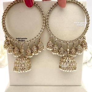 May include: A pair of gold-tone jhumka earrings with dangling pearl and rhinestone embellishments. The earrings feature a circular design with a large, ornate pendant at the bottom. The text "IndoExquisite Designs" is visible on the earrings.