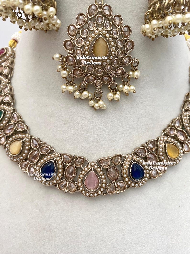 May include: An ornate, antique-style jewelry set featuring a necklace, earrings, and a headpiece. The set is silver-toned with a variety of colorful gemstones, including blue, pink, and yellow, and is accented with small pearls. The headpiece has a central teardrop-shaped stone.