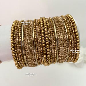 Peut inclure: Une pile de bracelets dorés avec diverses textures, notamment de petites perles et des embellissements scintillants. Les bracelets sont présentés sur une surface blanche. Le texte "IndoExquisite Designs" est visible.
