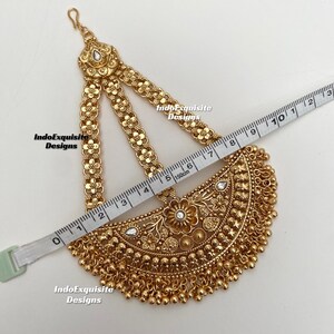 May include: A gold-toned headpiece featuring a semi-circular pendant with floral engravings, clear stones, and dangling beads. Three chains with flower motifs connect to a top hook. A measuring tape shows the piece is approximately 8.5 inches wide.