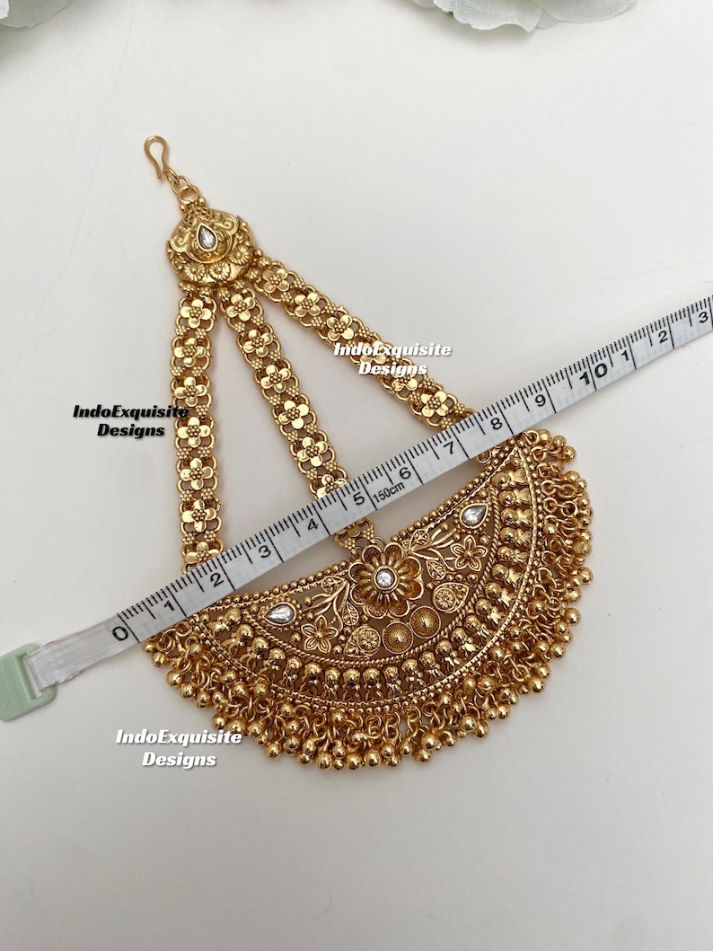 May include: A gold-toned headpiece featuring a semi-circular pendant with floral engravings, clear stones, and dangling beads. Three chains with flower motifs connect to a top hook. A measuring tape shows the piece is approximately 8.5 inches wide.
