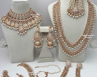 May include: A bridal jewelry set featuring a gold and pearl necklace, earrings, a maang tikka, a nose ring, a hathphool, and a waist chain. The set is decorated with white pearls and clear stones.
