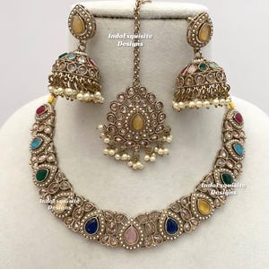 May include: A gold-toned jewelry set featuring a necklace, earrings, and a headpiece. The necklace and earrings have colorful gemstones and small pearls. The headpiece is a central pendant with a chain. The text "IndoExquisite Designs" is visible.