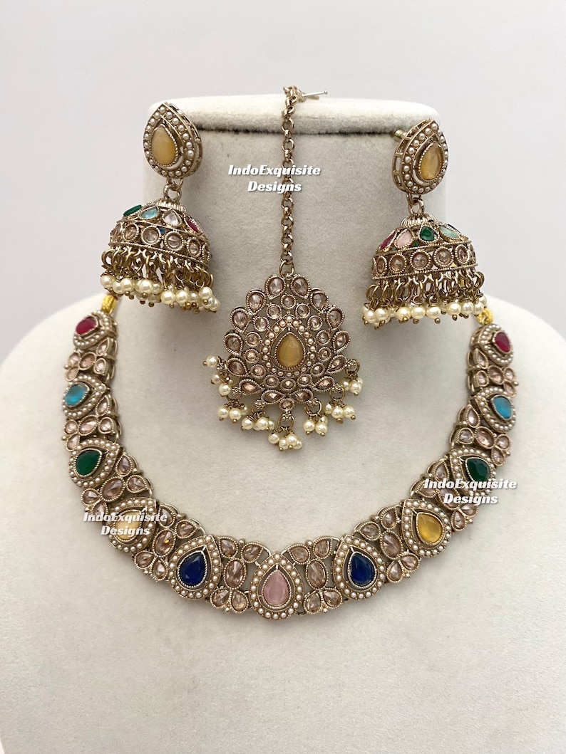 May include: A gold-toned jewelry set featuring a necklace, earrings, and a headpiece. The necklace and earrings have colorful gemstones and small pearls. The headpiece is a central pendant with a chain. The text "IndoExquisite Designs" is visible.