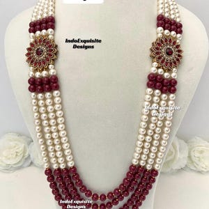 May include: A multi-strand necklace featuring alternating rows of white and burgundy beads. The necklace has a floral pendant with red gemstones and gold-colored accents. The text "Design 2" and "IndoExquisite Designs" are visible.