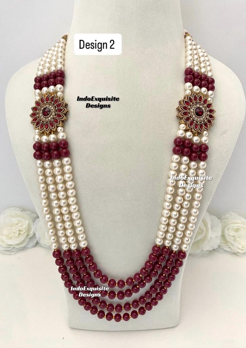 May include: A multi-strand necklace featuring alternating rows of white and burgundy beads. The necklace has a floral pendant with red gemstones and gold-colored accents. The text "Design 2" and "IndoExquisite Designs" are visible.