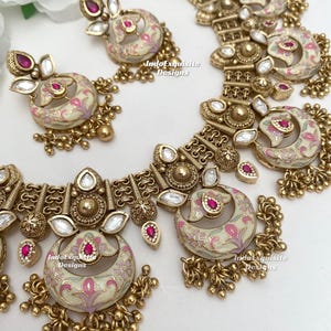 Pu&ograve; includere: Un set di gioielli color oro composto da collana e orecchini. Il set ha una base color crema con disegni floreali rosa e accenti rossi. La collana e gli orecchini hanno una forma a mezzaluna con perline dorate pendenti. Il testo "IndoExquisite Designs" &egrave; visibile.