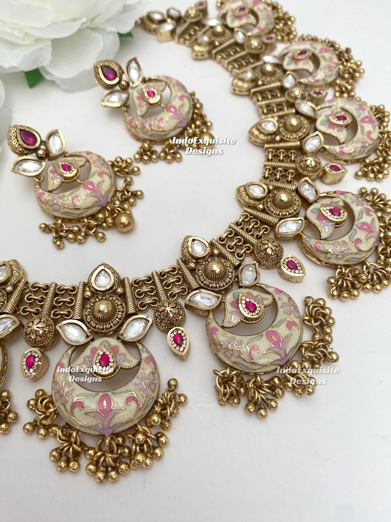 Pu&ograve; includere: Un set di gioielli color oro composto da collana e orecchini. Il set ha una base color crema con disegni floreali rosa e accenti rossi. La collana e gli orecchini hanno una forma a mezzaluna con perline dorate pendenti. Il testo "IndoExquisite Designs" &egrave; visibile.