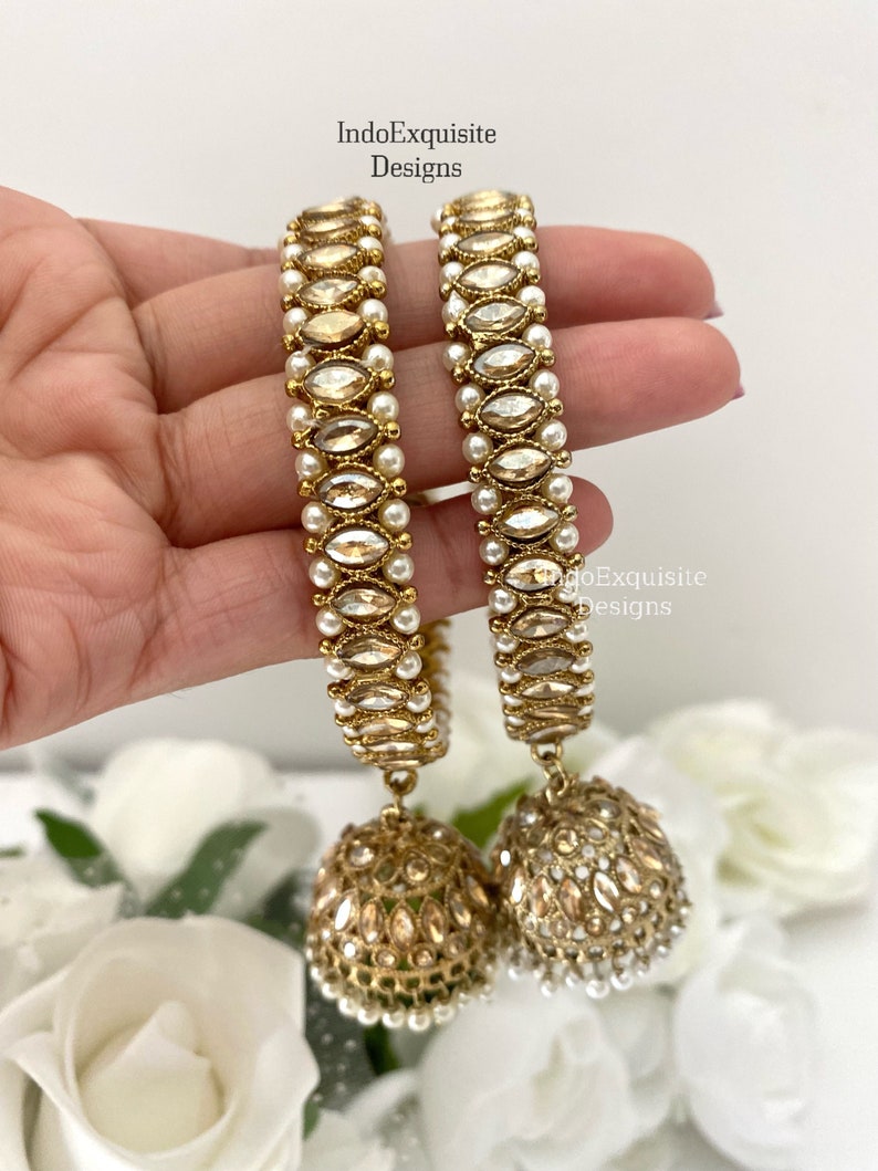May include: A pair of gold-tone jhumka earrings with white pearl accents and clear stones. The earrings have a dangling design with a round, ornate charm.