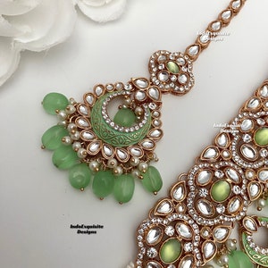May include: A gold and green Indian bridal headpiece with a large, ornate pendant and a chain of smaller pendants. The pendant features a green stone and is surrounded by clear rhinestones. The chain is adorned with green beads and pearls.